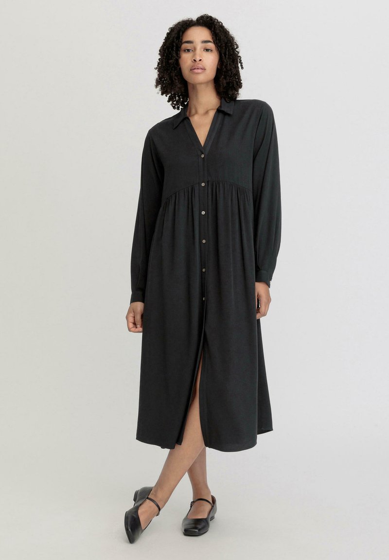 Schwarzes Langarmkleid mit durchgehender Knopfleiste, Kragen und Taillennaht. Es hat einen Schlitz vorne und eine glatte Stoffbeschreibung.