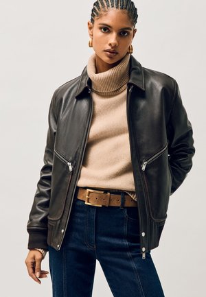 Kvinde med fletninger iført guldringformede øreringe, en sort læderjakke, beige rullekravetrøje, brun bælte og mørkeblå jeans.