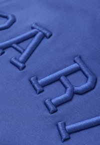 Bl ått tyg med upphöjda, satin-stickade bokstäver. Texturen är slät med en lätt glans, som framhäver de broderade elementen.