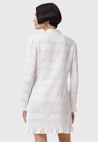 Robe blanche avec des rayures texturées verticales et un ourlet à volants. Manches longues, col, et subtile brillance du tissu. Vue de dos présentée.