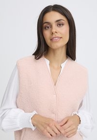 Világos rózsaszín gyapjú mellény, texturált anyagból, kerek nyakkal és gombos zárással, fehér blúzzal, fodros mandzsettákkal párosítva.