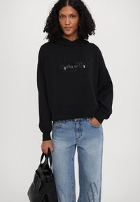Black hooded sweatshirt with a textured logo on the front, paired with light blue wide-leg jeans featuring a subtle pattern.