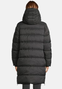 Femme portant un long manteau d'hiver matelassé noir à capuche, se tenant de dos contre un fond uni.