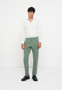 White long-sleeve shirt with button details, paired with light green tailored trousers and black shoes, set against a plain white background.