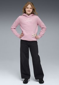 Rosa Kapuzenpullover mit weißem "Puma"-Logo, Fronttasche und dekorativen Akzenten. Kombiniert mit lockeren schwarzen Jeans und schwarzen Sneakers. Grauer Hintergrund.
