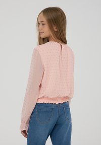 Blusa rosa chiaro con apertura a chiave sulla schiena, tessuto morbido, maniche lunghe, orlo elastico e motivo a pois dorati. Abbinata a jeans in denim blu.