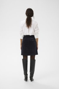 Sötétkék kordszoknya egyenes szabással, részletes hátsó zsebbel, fehér inggel és fekete térdfeletti csizmával viselve.