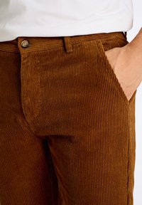 Pantalon en velours côtelé marron avec des côtes verticales, comportant une fermeture à bouton et une poche latérale. Le tissu présente une texture douce et un design durable.