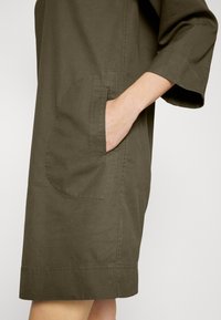 Main dans la grande poche avant d'une robe vert olive longueur genou avec des manches trois-quarts.