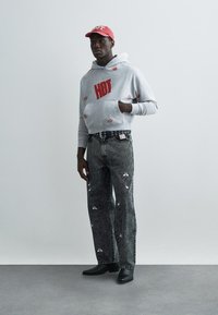 Grijze hoodie met rode "HOT" tekst en lipafdrukken, gecombineerd met gerafelde zwarte spijkerbroek met witte bloemenpatronen. Zwarte enkellaarzen. Rode pet.