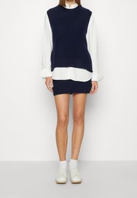 Top sin mangas tejido en color azul marino combinado con una camisa blanca de cuello y una falda corta azul marino. La modelo lleva calcetas blancas y zuecos con suelas de madera.