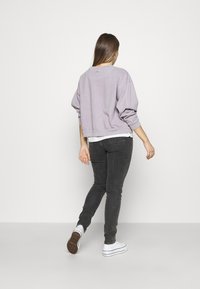 Sudadera de gran tamaño en color púrpura claro, con mangas largas; llevada sobre una camiseta blanca. Combinada con jeans skinny en gris oscuro y zapatillas blancas.