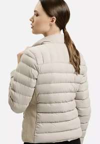Beżowa kurtka puchowa z pikowanymi sekcjami, wysokim kołnierzem oraz teksturowanymi, dzianinowymi bocznymi panelami. Dopasowany krój i lekka konstrukcja.