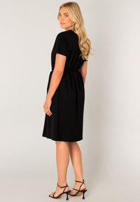 Zwart jurkje met korte mouwen, een getailleerde bovenkant en een cinched taille. De rok valt net boven de knieën en heeft een geplooid ontwerp. Gecoördineerd met zwarte hakken.