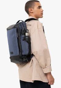 Zaino in rete blu e materiale nero, con tasca laterale per bottiglia d'acqua e cerniere, indossato su una spalla sopra una giacca beige.