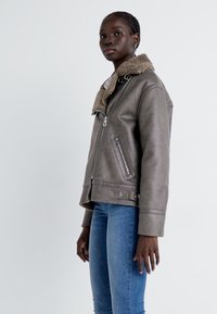 Grå detaljerad skinnjacka i faux läder med en shearlingkrage, dragkedja fram, sidofickor och en strukturerad passform. Buren över blå jeans.
