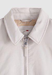 Gros plan sur une veste beige clair avec un col rembourré et une fermeture éclair argentée partiellement fermée, montrant une doublure intérieure beige.