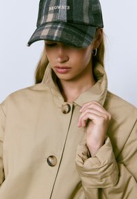 Chaqueta beige con un alto cuello, que presenta dos grandes botones. Una gorra de cuadros verde oscura con el texto "BROWNIE" se encuentra en la cabeza del modelo.