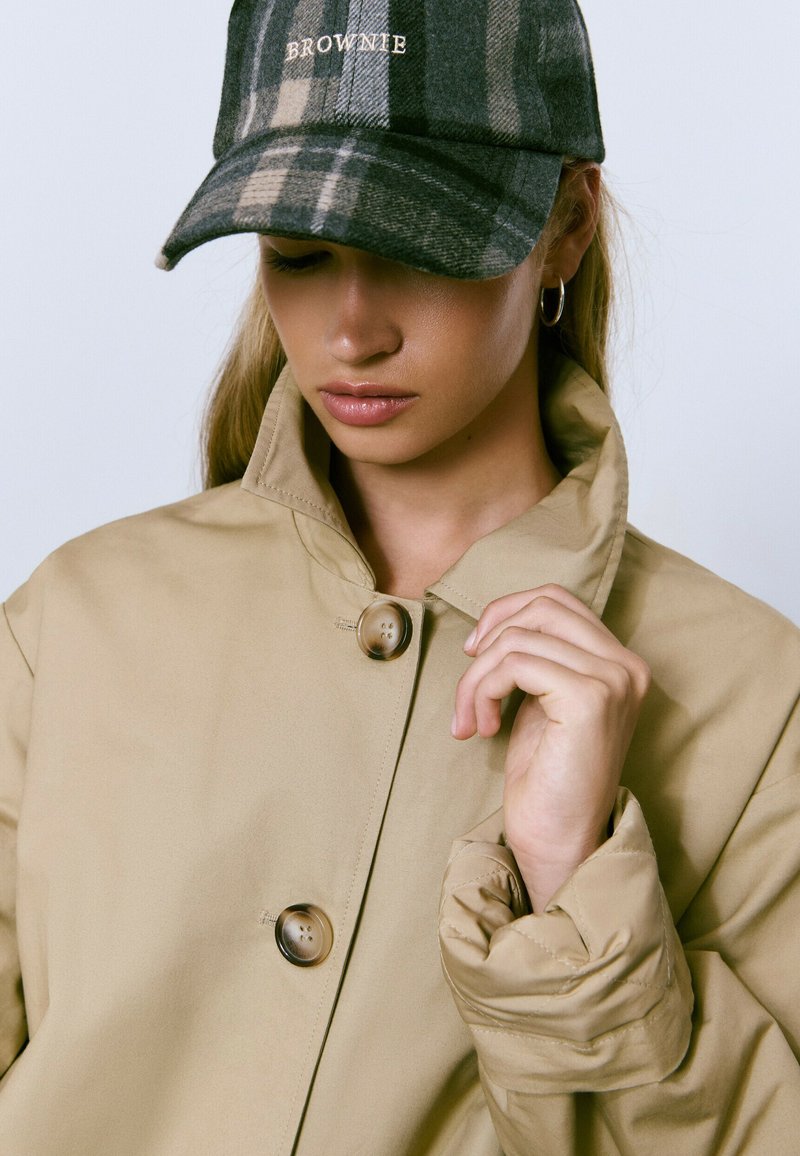 Chaqueta beige con un alto cuello, que presenta dos grandes botones. Una gorra de cuadros verde oscura con el texto "BROWNIE" se encuentra en la cabeza del modelo.
