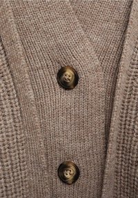 Cardigan tricoté en laine beige douce avec un motif texturé, doté d'un col en V et de deux boutons ronds marron foncé.