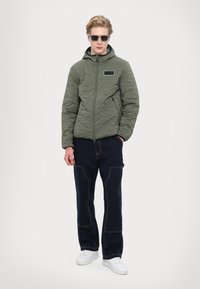 Quilted olive green hooded jacket with zippered pockets, paired with dark blue pants featuring visible stitching, and white sneakers.