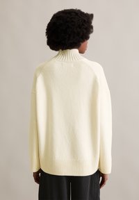 Ανοιχτόχρωμο πουλόβερ με ψηλό ραμμένο γιακά, πεσμένους ώμους και ραμμένο στρίφωμα. Μαλακή υφή, χαλαρή εφαρμογή και διακριτό σχέδιο πλέξης.