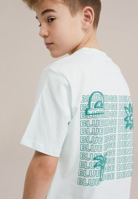 Minta zelena majica s kratkimi rokavi, ki vsebuje zloženo besedilo "BLUERIDGE DENIM" in grafiko palme na hrbtni strani. Mehak bombažni material.