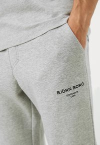 Harmaat collegehousut, jotka on valmistettu pehmeästä kankaasta ja joissa on sivutaskut. Mukana musta brändilogo vasemmassa reidessä ja rento istuvuus.