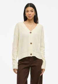 Gilet en tricot couleur crème avec un col en V, comportant cinq boutons, des manches longues et des poignets côtelés. Porté par-dessus un pantalon marron.