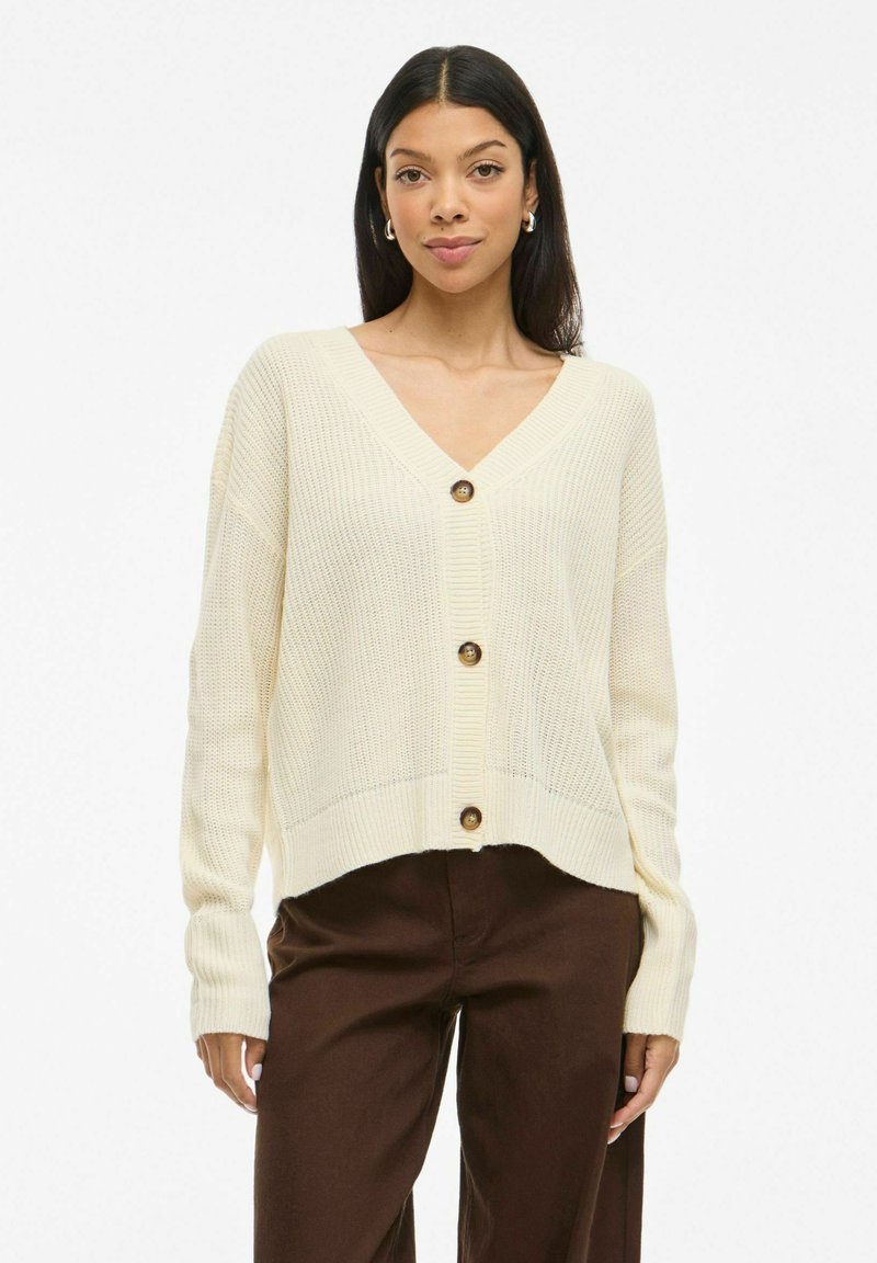 Gilet en tricot couleur crème avec un col en V, comportant cinq boutons, des manches longues et des poignets côtelés. Porté par-dessus un pantalon marron.