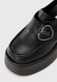 Melna ādas kurpe ar masīvu zoli, apjalbota ar sirds formas metāla sprādzi un gludu tekstūru. Elegants, noapaļots purngalu dizains.