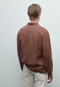 Chemise en lin marron avec un empiècement au dos, manches longues et poignets à boutons, associée à un pantalon gris clair. Texture lisse, coupe décontractée.