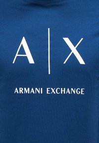 Μπλε πουκάμισο με λευκό λογότυπο "A | X" και το κείμενο "ARMANI EXCHANGE" στο κέντρο κάτω από το λογότυπο.