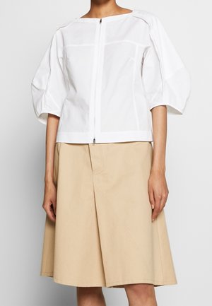 Blouse blanche avec fermeture éclair à l'avant et manches bouffantes trois-quarts, associée à une jupe beige trapèze longueur genou.