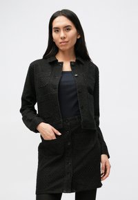 Čierna textúrovaná bunda s predným zapínaním na gombíky, dvoma náprsnými vreckami a zladenou sukňou. Nosená cez priliehavý čierny top s hladkým povrchom.