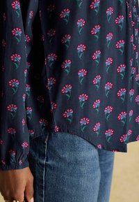 Tumši zili ziedu blūze ar maziem rozā un purpursarkaniem ziedu rakstiem, mīksta auduma, ar elastīgām aprocēm, apkopojot ar džinsiem jostasvietā.