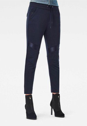 Pantalon bleu marine tapered avec poches latérales et détails de panneaux diagonaux, associé à des bottines noires à talons hauts avec fermetures éclair à l'avant.
