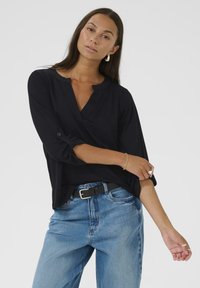 Blouse noire, à manches longues, légère, avec un col en V et des poignets retroussés. Associée à un jean en denim bleu clair taille haute et une ceinture noire.