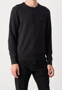 Maglione girocollo in cotone nero con polsini e orlo a costine, featuring un piccolo logo colorato sul petto. Texture liscia e vestibilità classica.