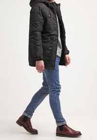 Svart vadderad jacka med huva, grå sweatshirt under, blå slim-fit jeans och vinröda Chelsea-boots med elastiska paneler.