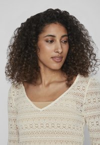 Kremfarget bluse med lange ermer i blonder, med V-hals og intrikate blomsterdesign, lagd over en nude tanktop. Krøllete hår delvis synlig.