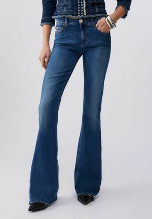 Donna con jeans scampanati blu scuro, scarpe nere appuntite e giacca di jeans con bottoni argentati, con una mano in tasca e braccialetti visibili.