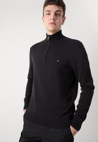 Sorte zip-neck genser i et fint strikket stoff. Har ribbestrikkede mansjetter og kant, med et lite logoemblem på venstre bryst.