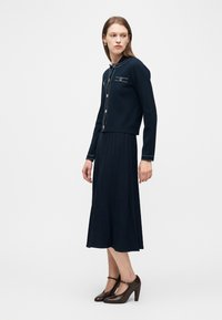 Γυναίκα που φοράει μπλε ναυτικό ζακέτα με διακοσμητικά κουμπιά και αντιστοιχισμένη πλισέ midi φούστα, σε συνδυασμό με καφέ ψηλοτάκουνα παπούτσια Mary Jane.