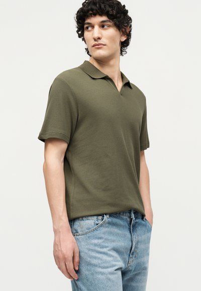 Jeune homme aux cheveux bouclés portant un polo manches courtes vert olive et un jean bleu clair, debout les mains dans les poches.