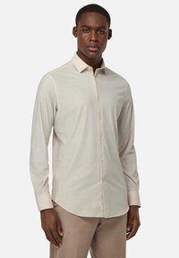 Camisa de manga larga beige clara con cuello abotonado, que presenta una textura de tela suave y un sutil detalle de costura a lo largo de las costuras.