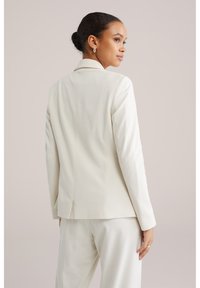 Kreemikollane kohandatud bleiser, millel on üks lõhike, kitsas lõige, sile tekstuur ja struktureeritud kraed. Kantud sobivate pükste kohal.