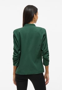 Grønn skreddersydd blazer med høy krage, rynkede ermer og en glatt tekstur, sett fra baksiden.