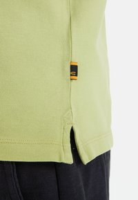 Lichtgroen katoenen polo-shirt met een gestructureerde stof; voorzien van zijsplitten en een klein zwart label met oranje accenten aan de onderrand.