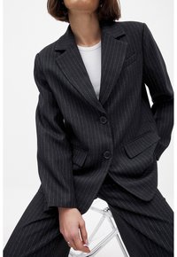 Femme assise sur une chaise transparente, vêtue d'un costume noir à fines rayures et d'un haut blanc, la main reposant sur la cuisse et la tête légèrement inclinée en arrière.